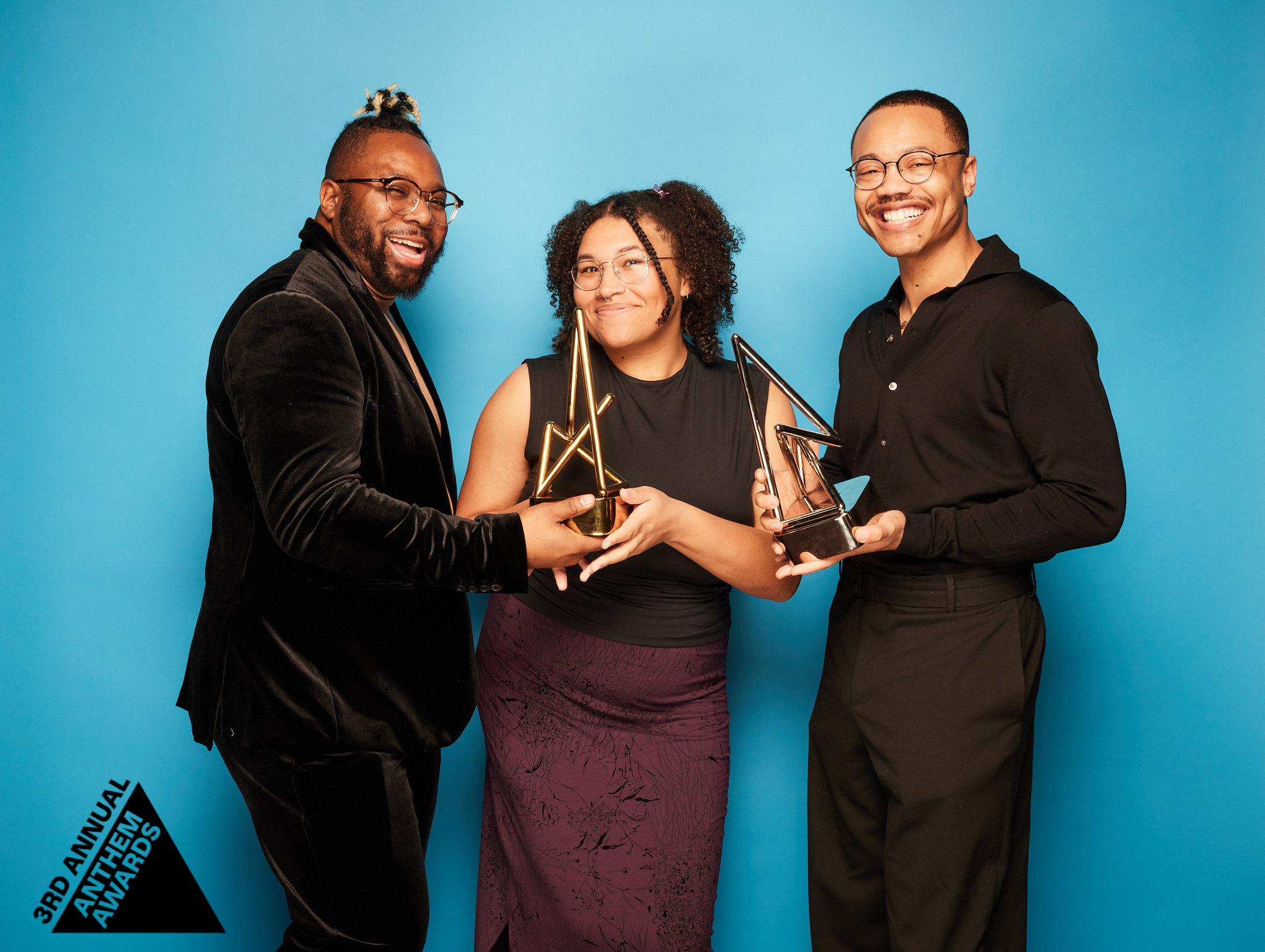 Text description: Three representatives from GLAAD at the 3rd Annual Anthem Awards. They are wearing black suits, holding a trophy in front of a blue backdrop.