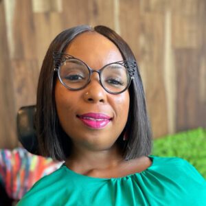 Smiling African-American woman wearing black turquoise and purple cat-eye glasses. She is wearing a black dress with red, purple, peach and turquoise flowers on it as well as peach floral earrings and red lipstick. Her hair is chin length natural curls. Although not depicted, she's a power wheelchair user.