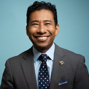 Image description: Franscisco Tezen wearing a gray suit and light blue shirt in front of a blue-grey background. He is smiling.