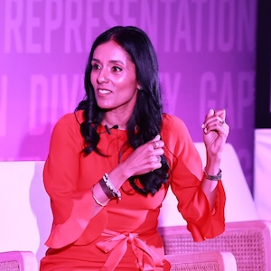 Image description: Woman named Mita Mallick in a red dress, speaking on a panel. She is sitting on a white chair, in front of a screen that reads Representation Diversity Gap.