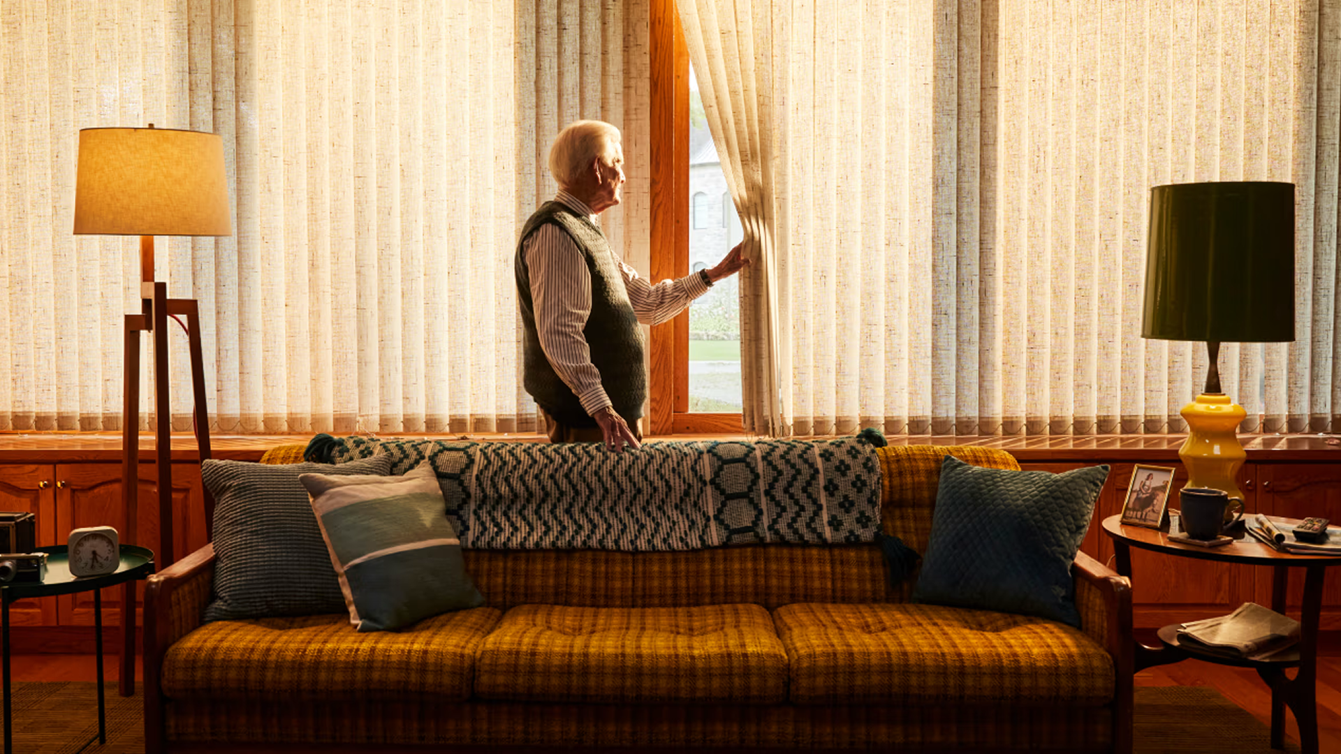 An older adult stands behind his couch in the living room, looking outside the window by peeling the curtain back.